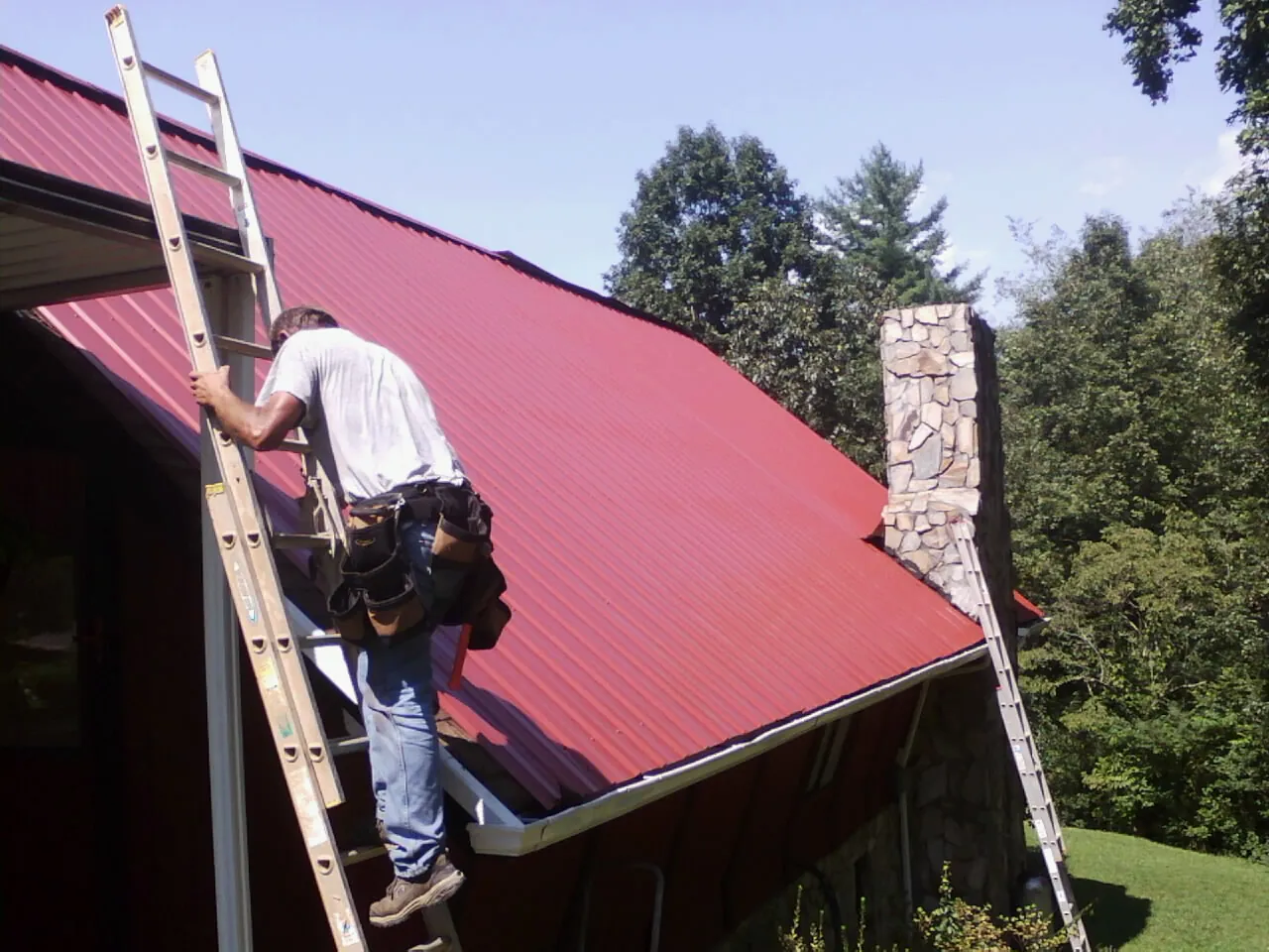 Roofer on a ladder beside a red metal roof and stone chimney — Soffit & Fascia Repair by Roofing Brothers in Oneco
